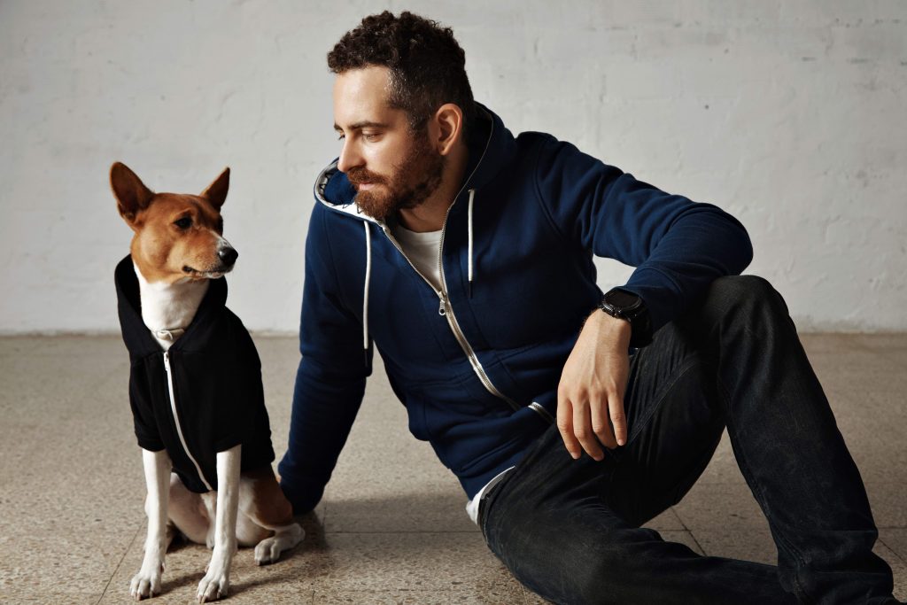 An attractive young man and his bassenji dog wearing matching hoodies sit on the floor next to white wall
